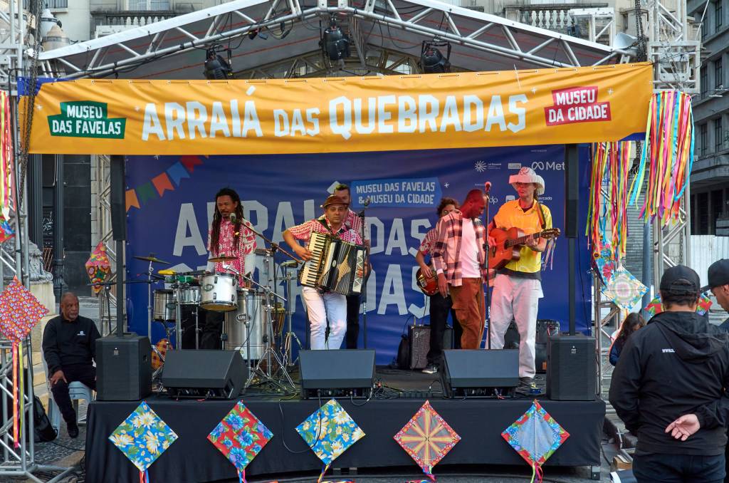 Banda de forró tocando em palco de rua decorado com bandeirinhas e fitas coloridas. Um sanfoneiro, um violonista de chapéu de palha, um baterista de dreadlocks e um baixista se apresentam sob uma faixa amarela com os dizeres Arraiá das Quebradas. Um homem de camisa preta observa sentado à esquerda, e pessoas assistem de costas à direita