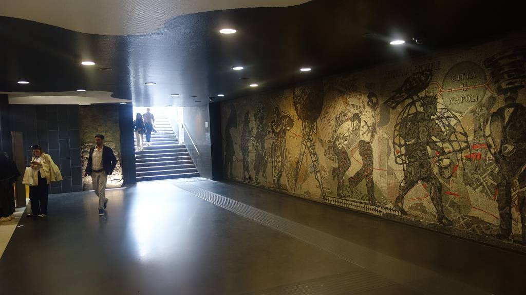 Pessoas em um corredor subterrâneo com teto escuro e iluminação embutida. À direita, um grande mosaico colorido com figuras humanas e a inscrição CITTÀ DI NAPOLI. À esquerda, uma escadaria iluminada leva a uma saída. Um homem de blazer escuro e calça clara caminha no centro, enquanto outras pessoas sobem a escada e uma mulher de casaco claro consulta o celular à esquerda