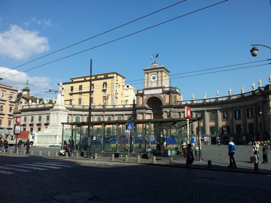 Praça com edifícios históricos em Nápoles, Itália, sob céu azul. À esquerda, estátua branca e prédio amarelo. Ao centro, estrutura com relógio e bandeiras. Pessoas caminham na praça e em uma parada de ônibus com grafites. O chão é de paralelepípedos. Foto sob licença CC BY-SA 3.0