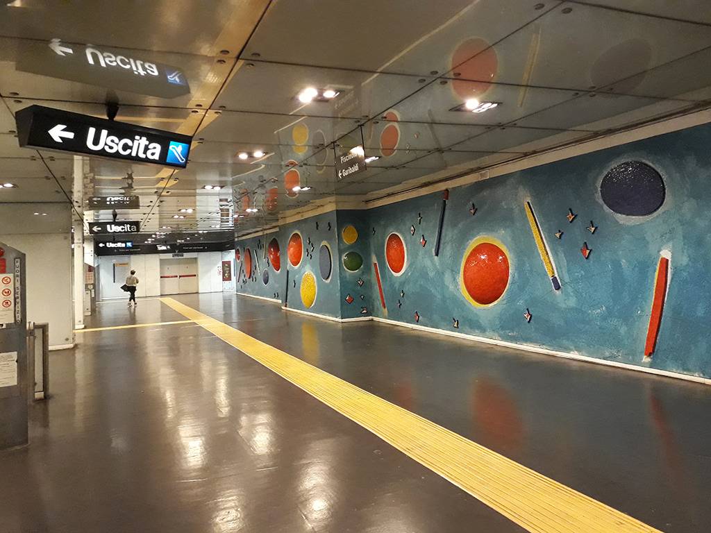 Corredor de metrô com piso escuro e faixa tátil amarela, paredes azuis decoradas com círculos coloridos e formas geométricas. Placas pretas indicam Uscita (Saída) com setas para a esquerda. Uma pessoa caminha ao fundo, à esquerda. Foto sob licença CC BY-SA 4.0