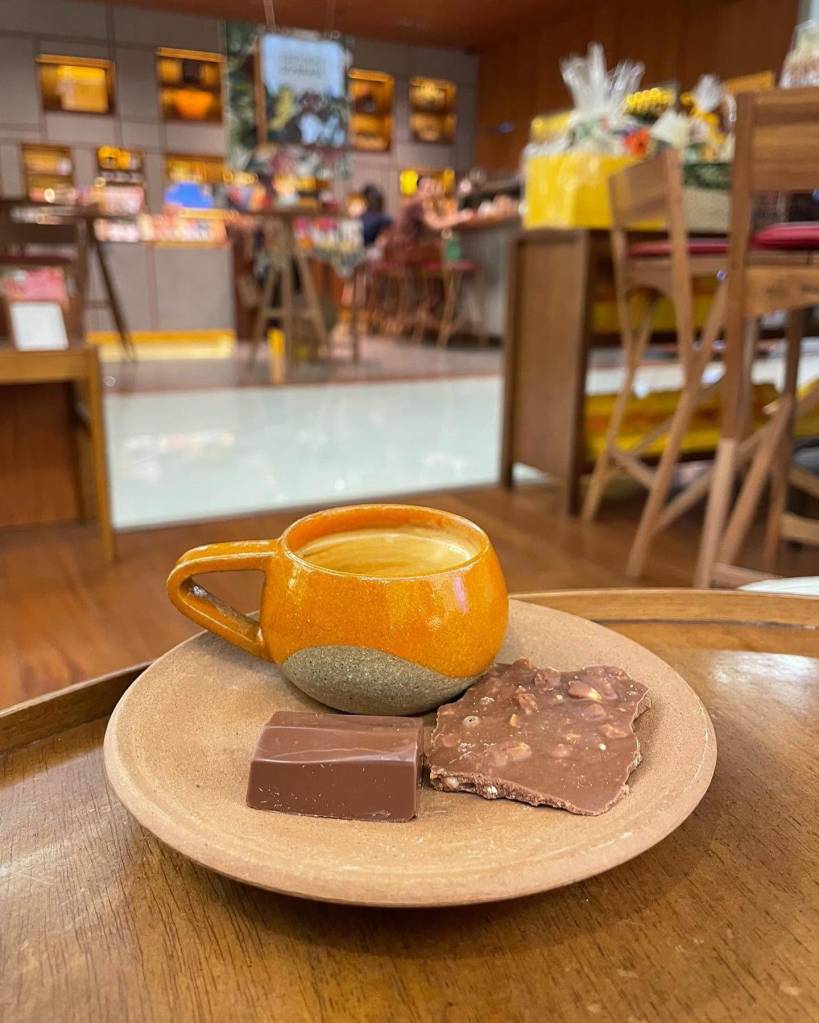 Xícara de café expresso em cerâmica laranja e cinza, com dois pedaços de chocolate, um liso e outro com castanhas, sobre um prato de cerâmica marrom em uma mesa de madeira. Ao fundo, um café borrado com pessoas e prateleiras.
