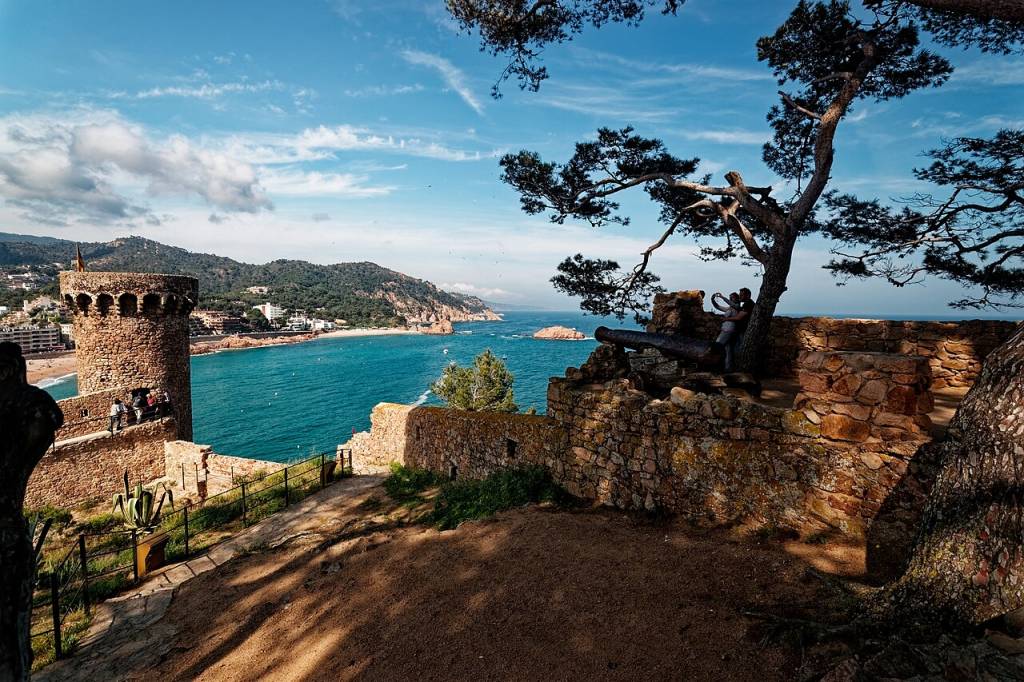 Vista panorâmica de uma torre de pedra antiga à esquerda, com pessoas no topo, e um canhão sobre um muro de pedra à direita, onde um casal se abraça sob uma árvore. O mar azul-turquesa e uma praia com montanhas ao fundo completam a paisagem sob um céu azul com nuvens esparsas. Foto sob licença CC BY-SA 4.0
