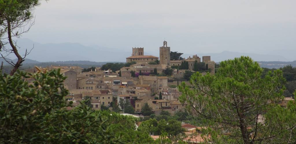 Vista panorâmica de um vilarejo medieval com casas de pedra e telhados terrosos, cercado por árvores verdes. Ao fundo, montanhas azuladas sob um céu nublado. Duas torres de castelo se destacam no centro da imagem. Foto sob licença CC BY-SA 3.0