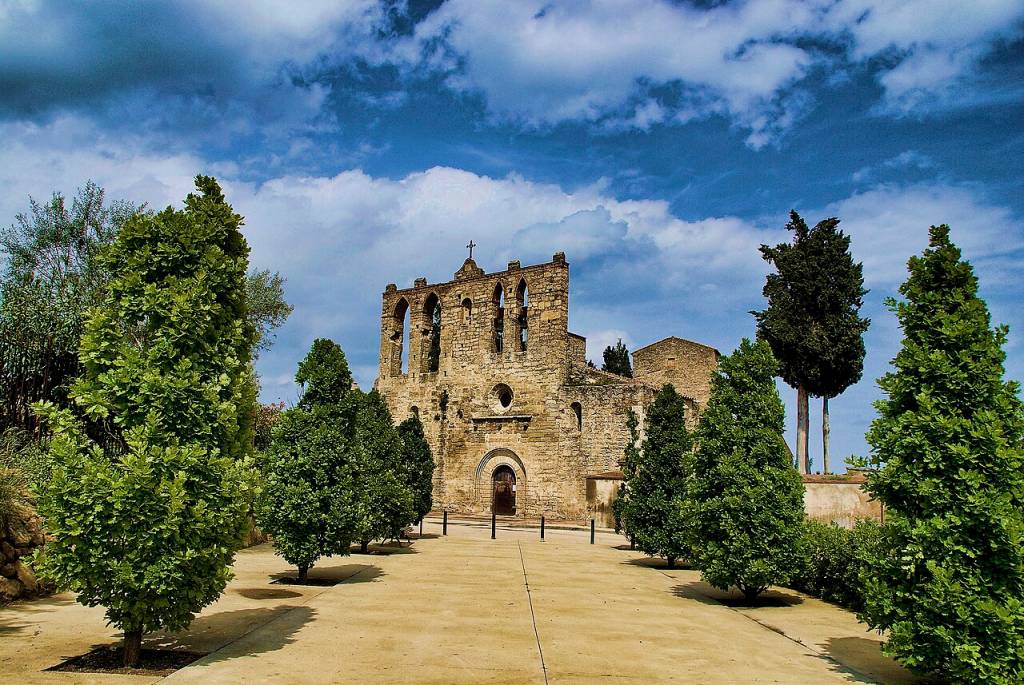 Antiga igreja de pedra com torre sineira em ruínas, com uma cruz no topo, em um dia ensolarado com céu azul e nuvens brancas. Uma alameda de terra clara ladeada por árvores verdes e arbustos leva à entrada principal da igreja, que tem um arco de pedra e uma porta de madeira escura. Ao fundo, outras construções de pedra e mais árvores.
