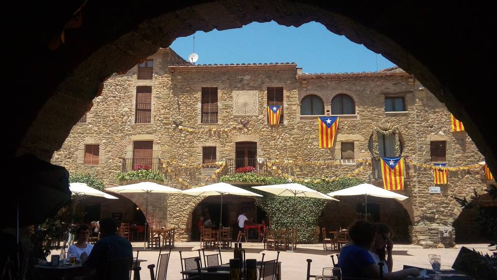Vista através de um arco escuro para uma praça ensolarada com edifícios de pedra antigos, decorados com bandeiras catalãs e um relógio de sol. Pessoas sentam-se em mesas sob guarda-sóis brancos, desfrutando do ambiente externo. Foto sob licença CC BY-SA 4.0