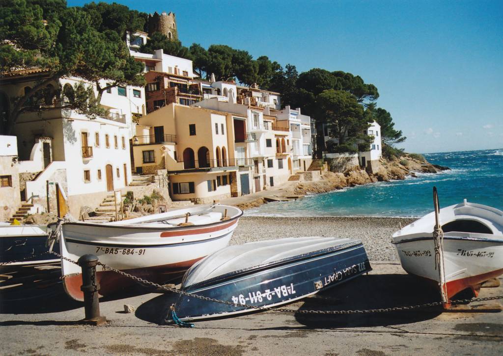 Três barcos de pesca em uma praia de pedras, com casas brancas e beges em uma encosta arborizada, e o mar azul-turquesa com ondas ao fundo. CC BY 3.0