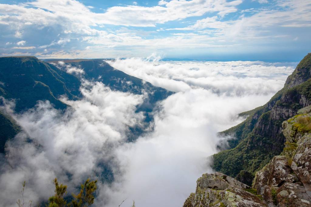 Cânion com vales profundos e encostas verdes, parcialmente cobertos por um denso mar de nuvens brancas, sob céu azul com nuvens esparsas