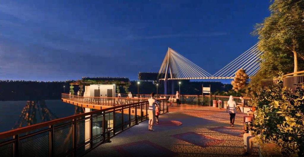 Pessoas em um calçadão iluminado à noite, com um rio à esquerda e uma ponte estaiada ao fundo. O céu é azul escuro, e árvores frondosas margeiam o lado direito do calçadão.