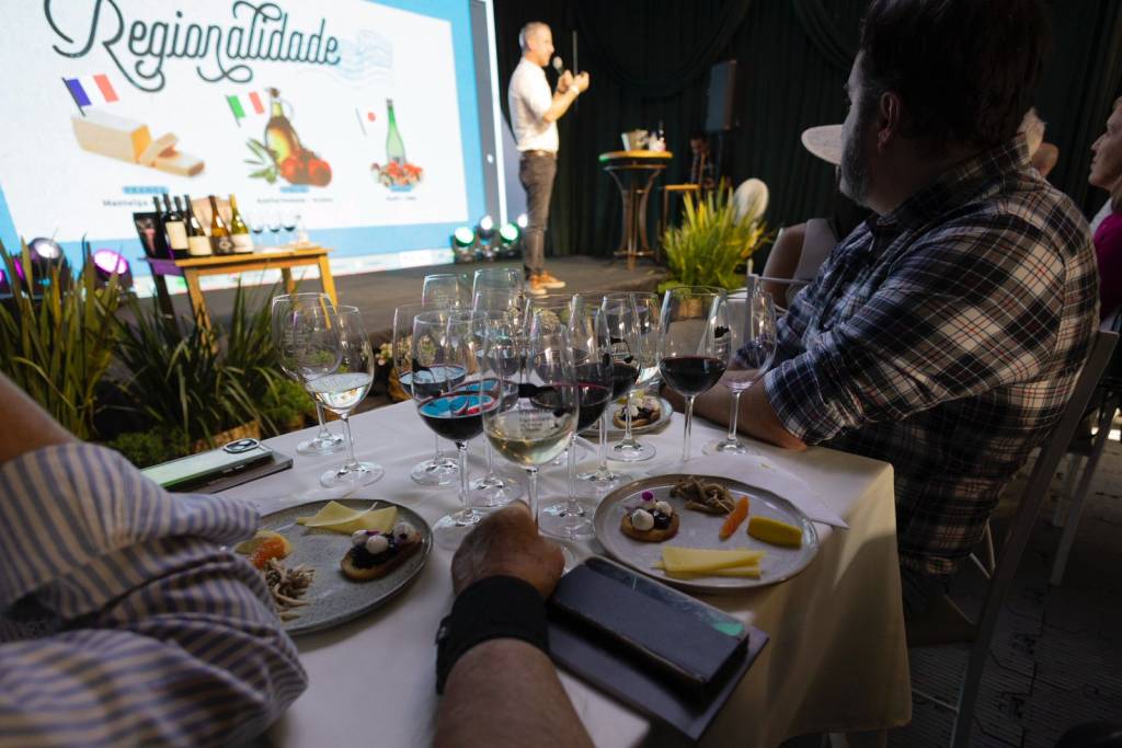 Pessoas em uma degustação de vinhos, com taças de vinho tinto e branco sobre a mesa, acompanhadas de pratos com queijos e petiscos. Ao fundo, um homem apresenta um telão com o título Regionalidade e imagens de produtos típicos de diferentes países