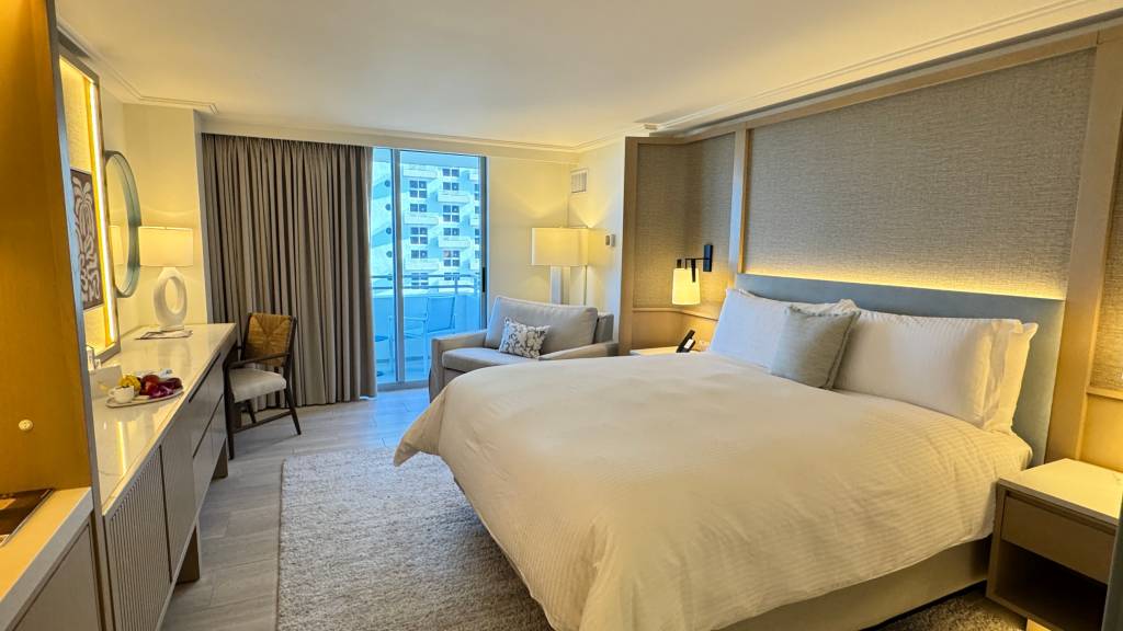 Quarto de hotel luxuoso com cama king-size de lençóis brancos, cabeceira estofada e iluminação indireta. À direita, uma mesa de cabeceira com luminária. À esquerda, um sofá cinza, uma poltrona de madeira e uma bancada com espelho, fruteira e luminária. Ao fundo, uma porta de vidro dá para uma varanda com vista para prédios. O ambiente tem piso claro e carpete felpudo.