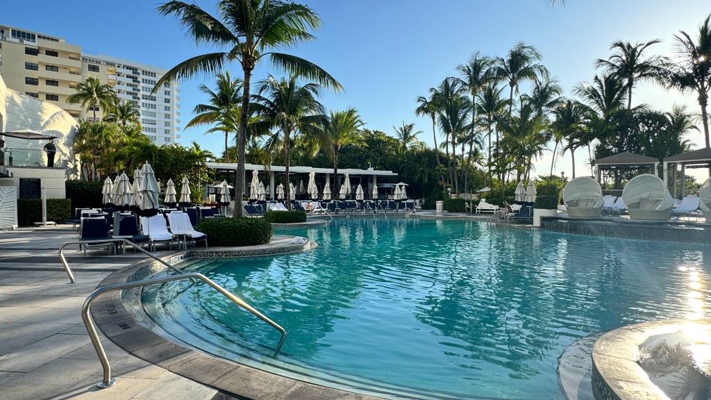 Piscina de hotel com água azul-turquesa, rodeada por espreguiçadeiras, guarda-sóis brancos e palmeiras. Ao fundo, prédios altos e o sol refletindo na água, criando um ambiente de lazer e relaxamento. Um homem observa da varanda à esquerda