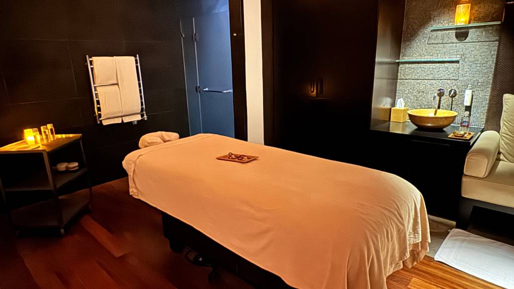 Sala de spa com iluminação suave, maca de massagem coberta por lençol pêssego, óculos sobre bandeja de madeira. À esquerda, mesa de apoio com velas acesas e toalhas em aquecedor. À direita, pia dourada sobre balcão escuro e poltrona creme. O ambiente é relaxante e acolhedor