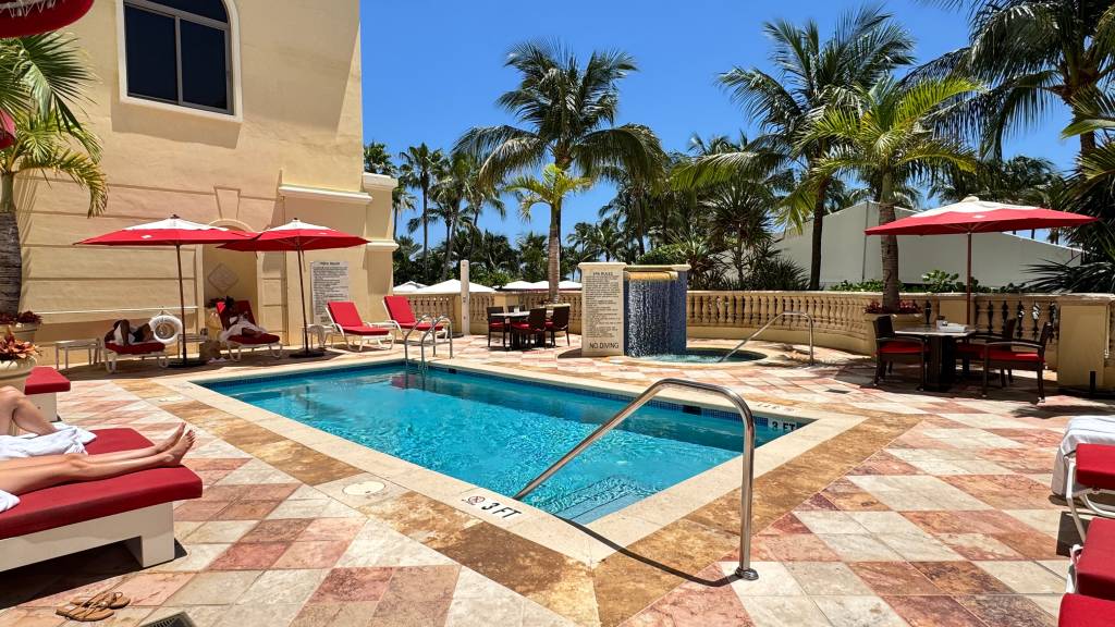 Piscina retangular de hotel com água azul, cercada por espreguiçadeiras vermelhas e guarda-sóis. Palmeiras altas e um prédio bege com janelas escuras ao fundo, sob céu azul claro. Uma pessoa relaxa em uma espreguiçadeira à esquerda.