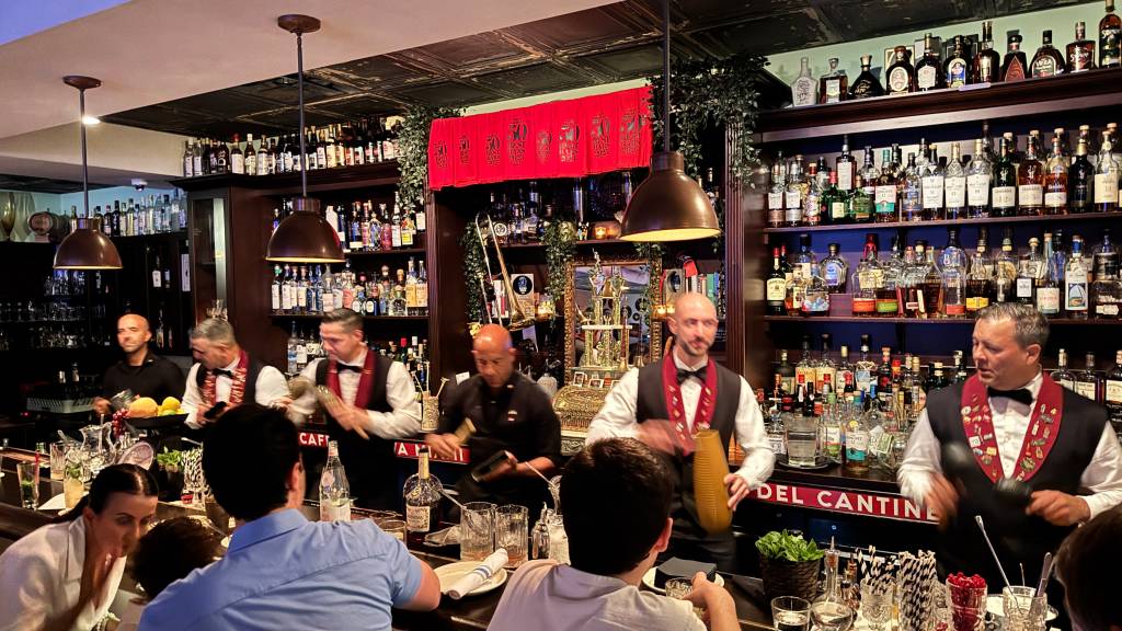 Seis bartenders, cinco homens e uma mulher, preparam bebidas em um bar com prateleiras cheias de garrafas de bebidas alcoólicas. Dois homens usam coletes pretos com laços vermelhos e medalhas, enquanto os outros usam camisas pretas. Clientes sentam-se à frente do balcão, observando a preparação. O ambiente é iluminado por pendentes de teto e decorado com folhagens e um espelho dourado. Uma faixa vermelha com caracteres dourados decora a parede.