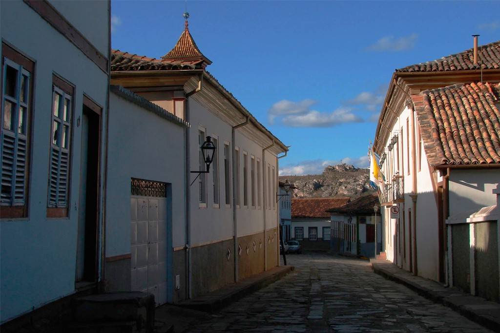 Rua de paralelepípedos em cidade histórica, ladeada por casarões coloniais de telhados de barro e paredes claras. À esquerda, uma torre pontiaguda no telhado. Ao fundo, montanhas sob céu azul com nuvens esparsas