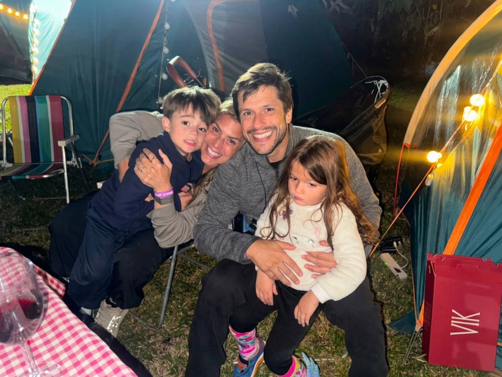 Família feliz acampando à noite, com pai, mãe e dois filhos pequenos sorrindo para a câmera, cercados por barracas iluminadas