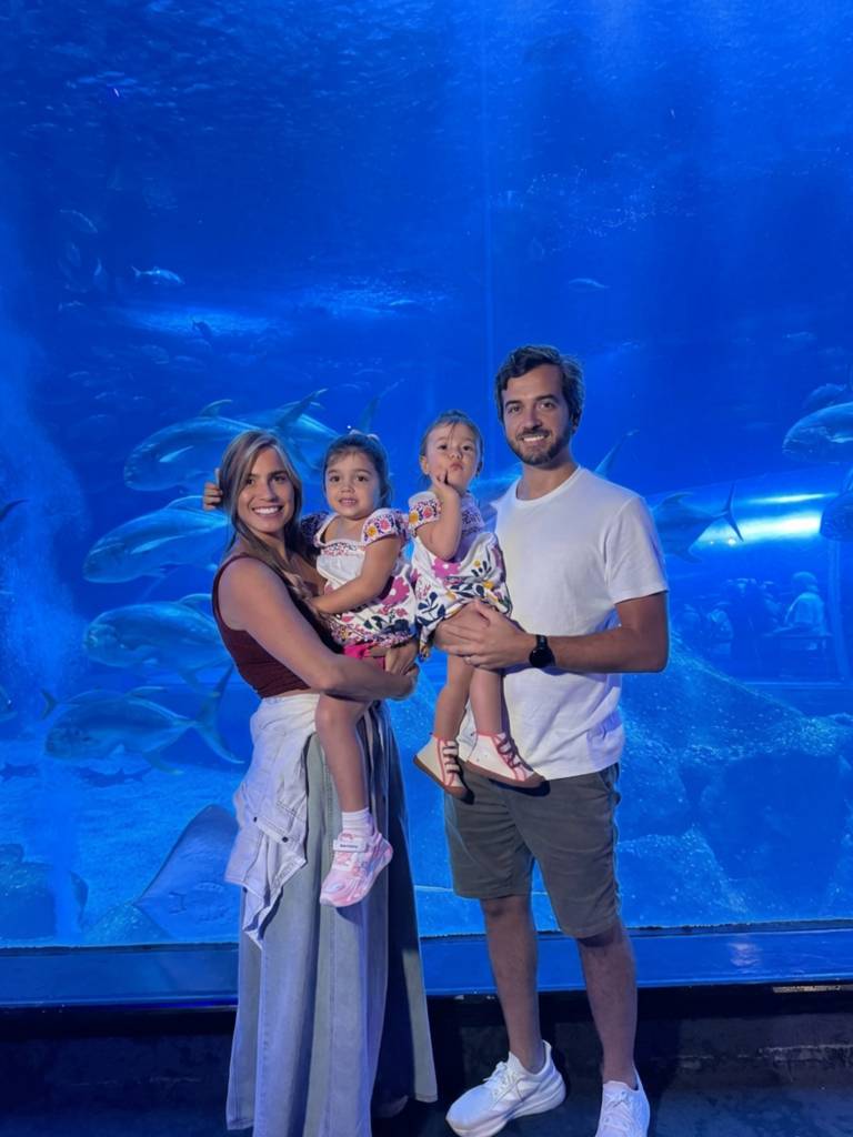 Família sorridente, pai, mãe e duas filhas pequenas, posando em frente a um grande aquário com tubarões e peixes nadando em água azul