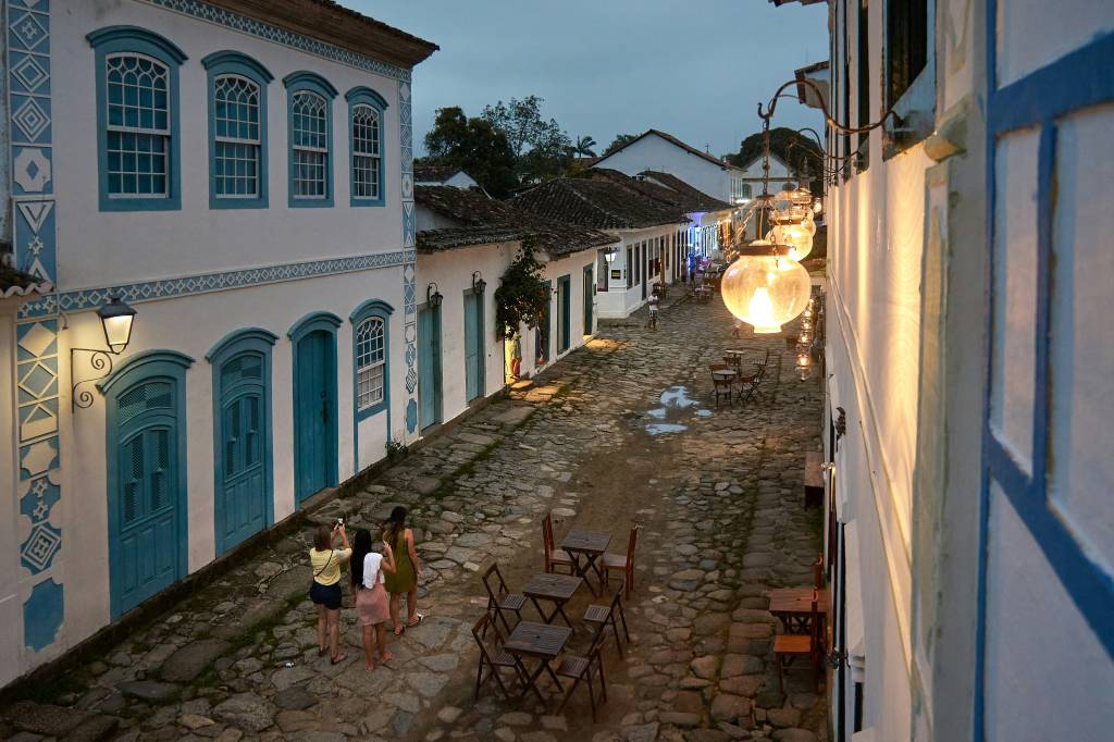 Vista aérea de uma rua de paralelepípedos em Paraty ao entardecer, com três mulheres caminhando e tirando fotos. As casas coloniais brancas têm portas e janelas azuis, e luminárias antigas iluminam o caminho, onde mesas de madeira estão dispostas em frente aos estabelecimentos