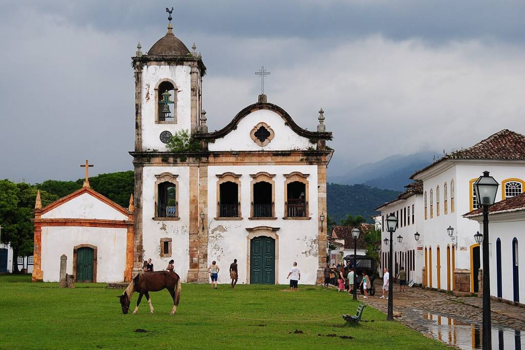 Igreja de Santa Rita em Paraty, com fachada branca e torre sineira. Um cavalo pasta na grama verde à frente, enquanto pessoas caminham pela praça e rua de pedra molhada. Casas coloniais brancas ladeiam a rua, e montanhas encobertas por nuvens escuras ao fundo. O céu está nublado