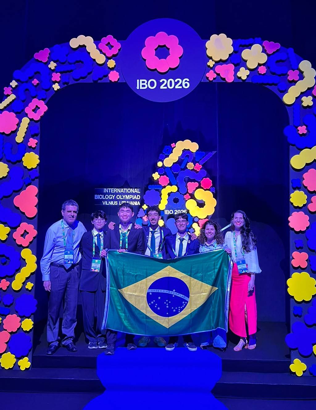 SI_butantan_site02 Seis jovens e dois adultos sorridentes posam com a bandeira do Brasil sob um arco decorado com flores coloridas e o letreiro IBO 2026
