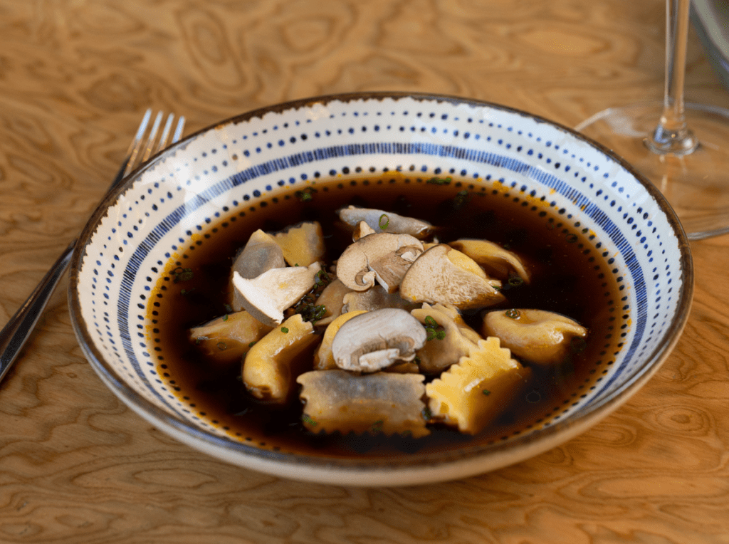 Uma tigela de cerâmica com borda azul e branca, cheia de caldo escuro, cogumelos fatiados e raviólis amarelos. Um garfo e um copo de vinho aparecem desfocados ao fundo, sobre uma mesa de madeira clara
