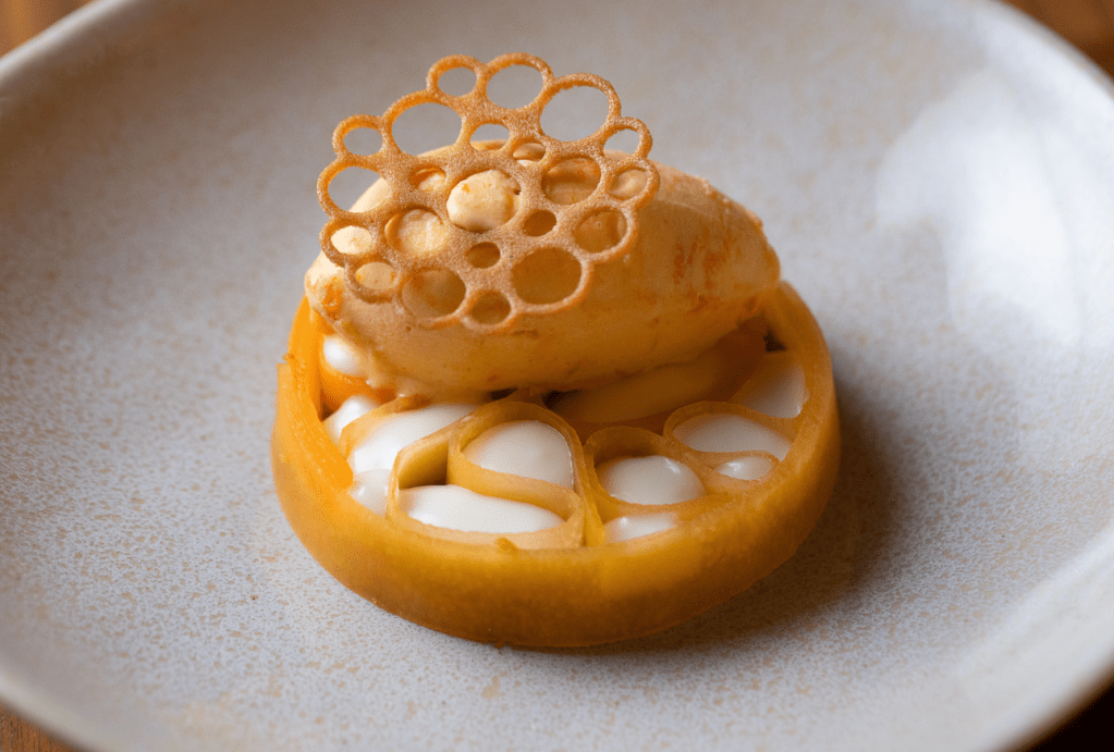 Sobremesa gourmet com sorvete laranja cremoso sobre uma base de fruta cortada, recheada com creme branco e decorada com um biscoito rendado em formato de flor de lótus, servida em prato claro
