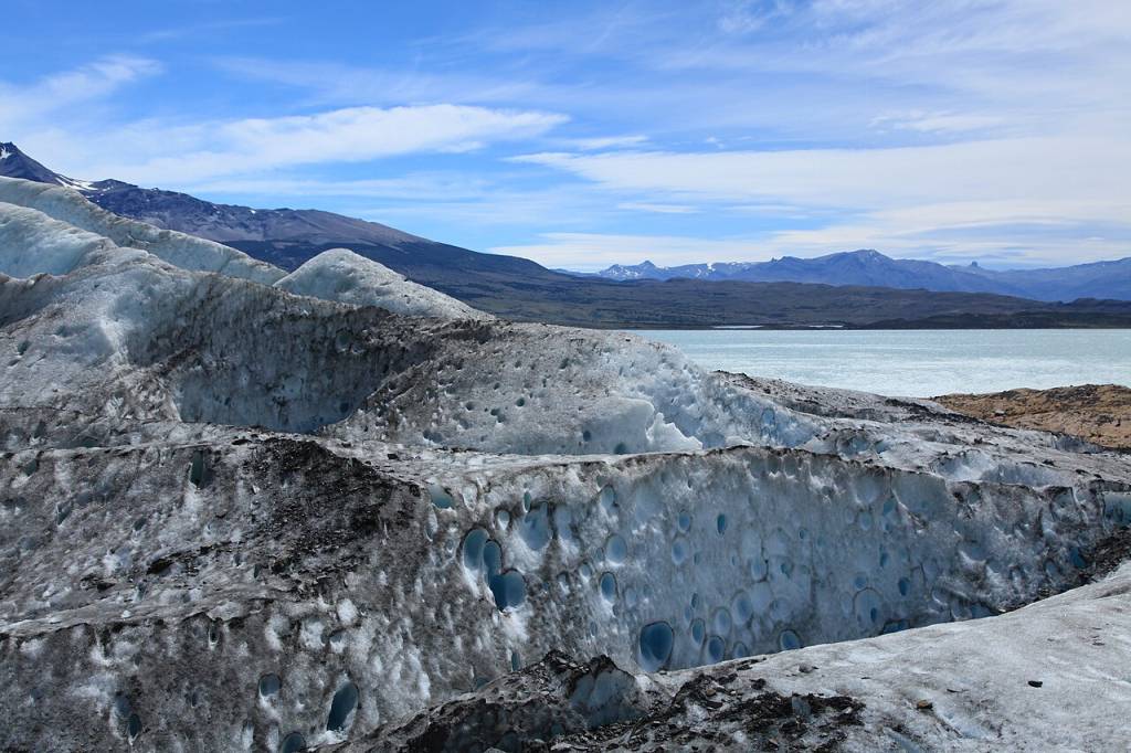 Geleira com gelo azul e cinza escuro, com buracos e fendas, à beira de um lago azul-claro. Montanhas escuras e picos nevados ao fundo, sob céu azul com nuvens esparsas