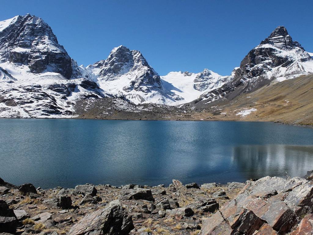 Lago azul-turquesa em primeiro plano, com margens rochosas e grama seca. Ao fundo, montanhas imponentes com picos nevados e encostas marrons sob um céu azul claro