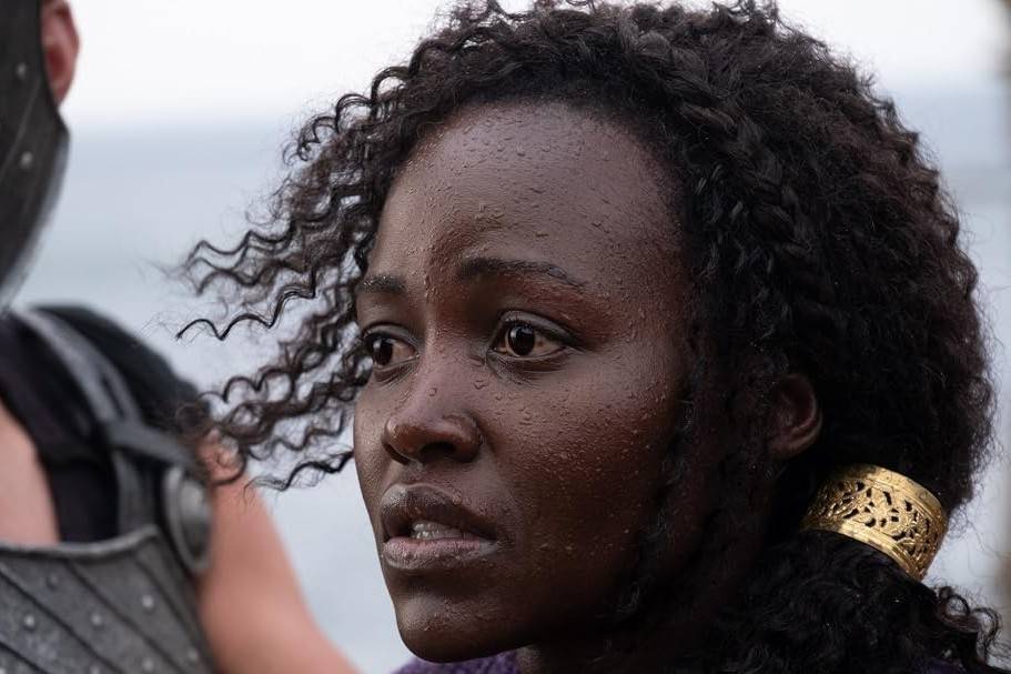 Mulher negra com cabelo crespo e molhado, usando um adorno dourado no cabelo, olha para o lado com expressão séria. Gotas de água cobrem seu rosto e cabelo. Ao fundo, um ombro com armadura e céu nublado