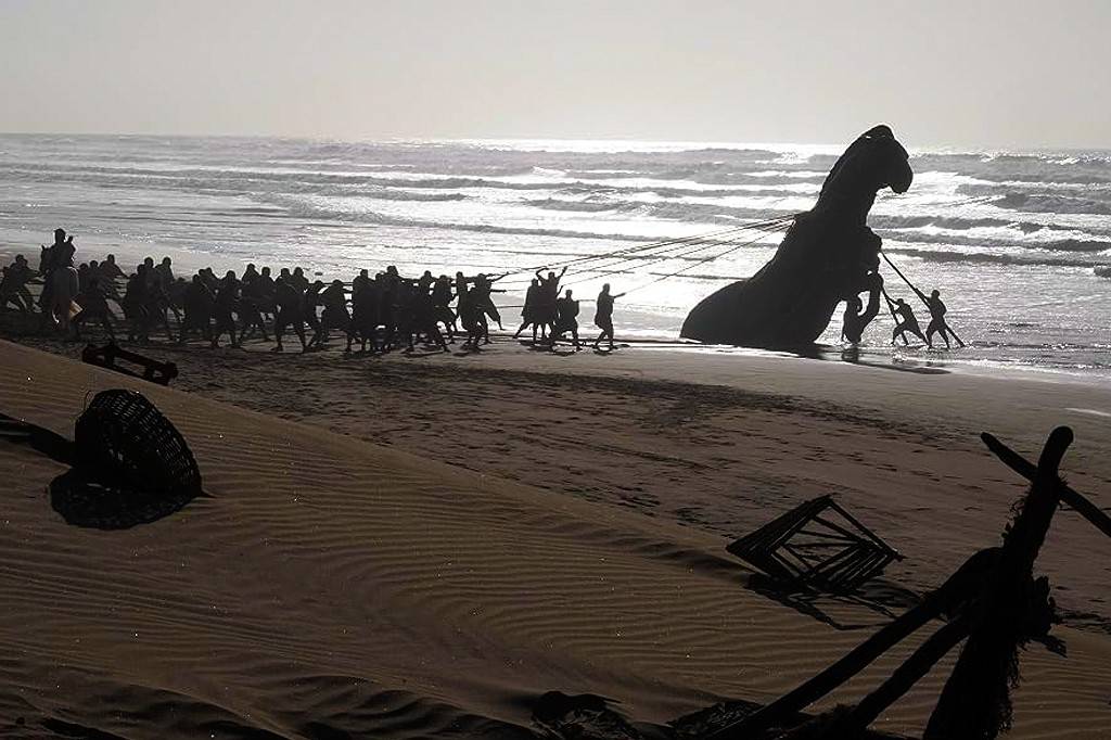 Silhuetas de dezenas de pessoas puxando cordas conectadas a uma grande estrutura escura em forma de cavalo, parcialmente submersa na água do mar, em uma praia arenosa com ondas ao fundo e céu claro