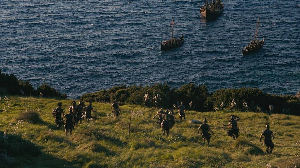 Guerreiros com armaduras escuras correm por uma colina gramada em direção a barcos antigos no mar azul, sob um céu claro