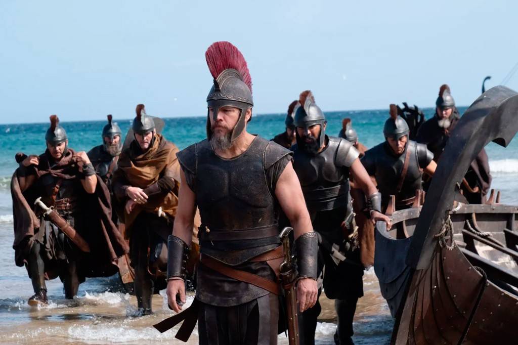Homens com armaduras e capacetes antigos, um com penacho vermelho, desembarcam de um barco de madeira na praia, com o mar azul ao fundo