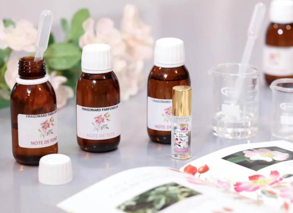 Frascos de perfume Fragonard em tons de âmbar e um frasco roll-on transparente, com pipetas e béqueres de vidro, sobre uma mesa com um livro aberto exibindo flores e frutos vermelhos