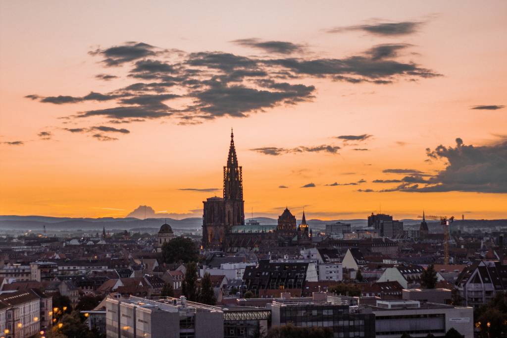 Vista aérea de uma cidade europeia ao pôr do sol, com o céu em tons de laranja e nuvens escuras. No centro, uma catedral gótica com torre alta e pontiaguda se destaca entre os telhados escuros das casas e edifícios. Montanhas ao fundo completam a paisagem urbana