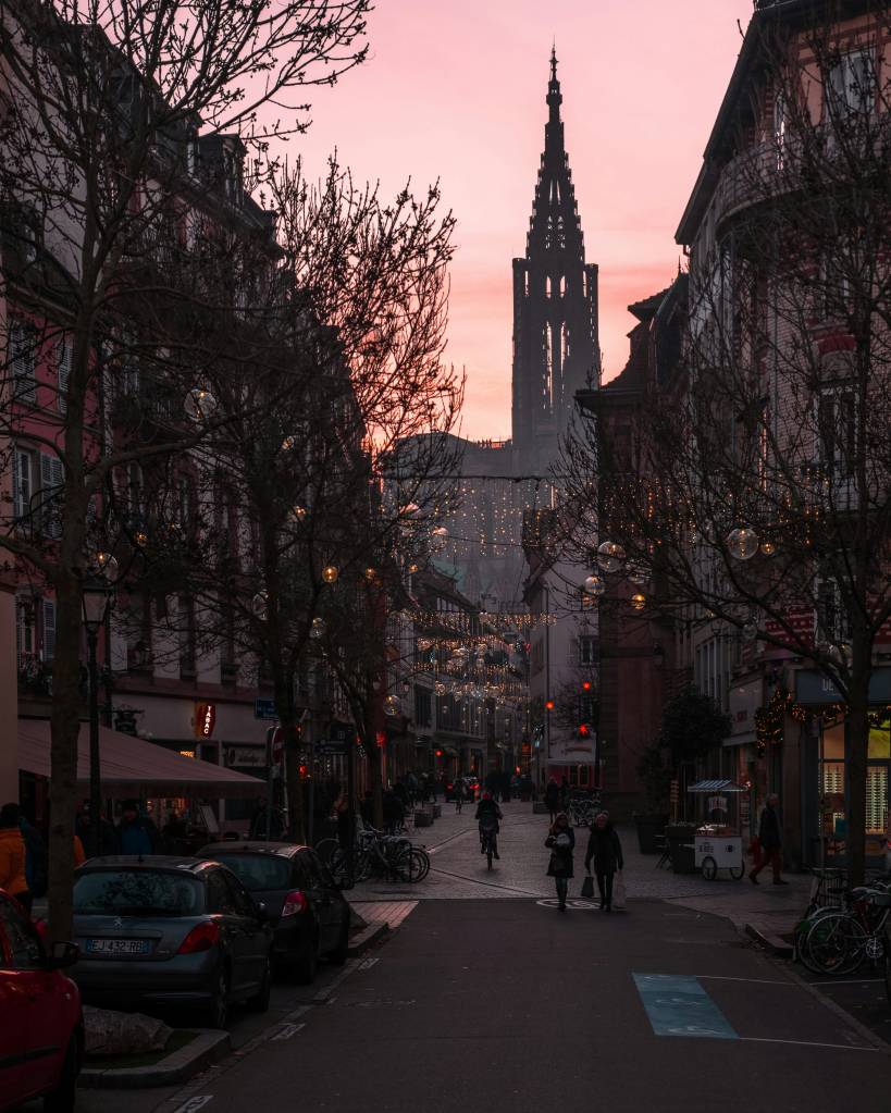 Rua estreita em cidade europeia ao entardecer, com céu rosa e torre de igreja gótica ao fundo. Árvores sem folhas com luzes decorativas e pessoas caminhando na calçada e na rua. Carros estacionados à esquerda e bicicletas à direita. Edifícios antigos ladeiam a rua, alguns com toldos e vitrines iluminadas
