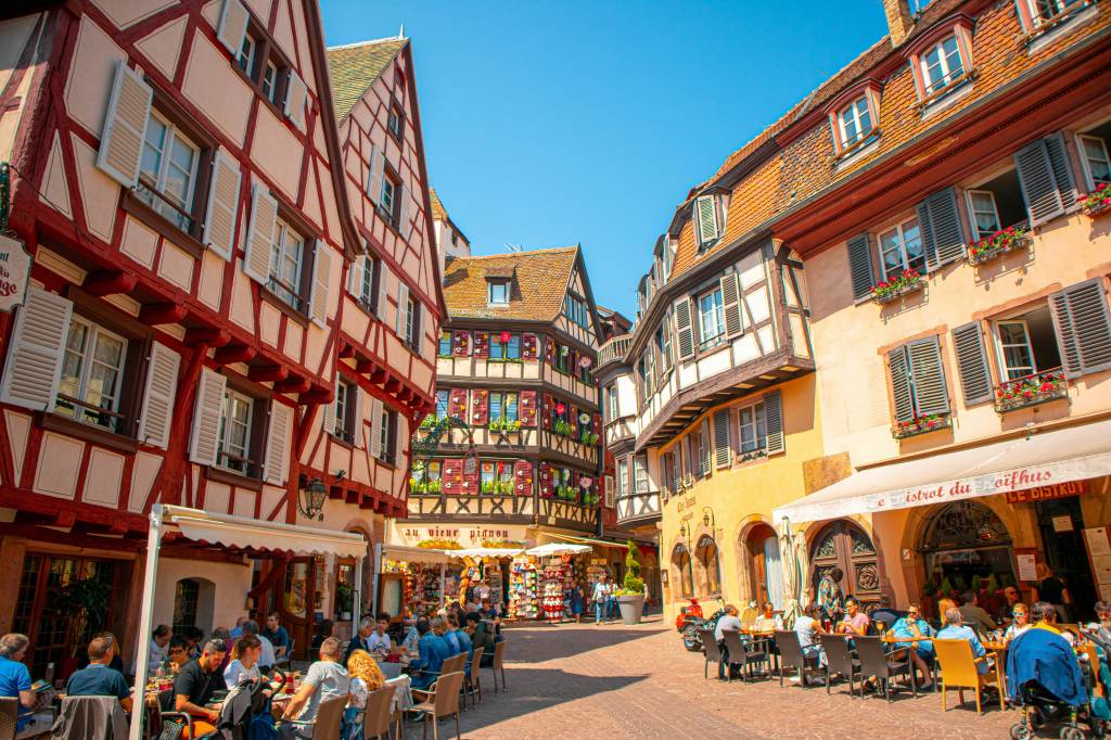 Rua movimentada em Colmar, França, com edifícios coloridos de arquitetura enxaimel e telhados inclinados. Pessoas sentadas em mesas de restaurantes ao ar livre, sob toldos brancos, desfrutando do dia ensolarado. Janelas com floreiras e persianas abertas adornam as fachadas.