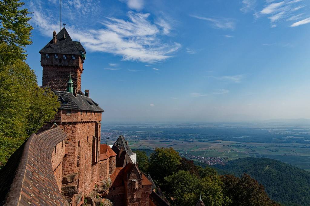 Castelo Haut-Kœnigsbourg, com paredes de tijolos avermelhados e telhados escuros, em uma montanha arborizada. O céu azul claro tem nuvens esparsas e, ao fundo, um vale vasto com campos e pequenas cidades se estende até o horizonte. Foto sob licença CC BY-SA 2.0