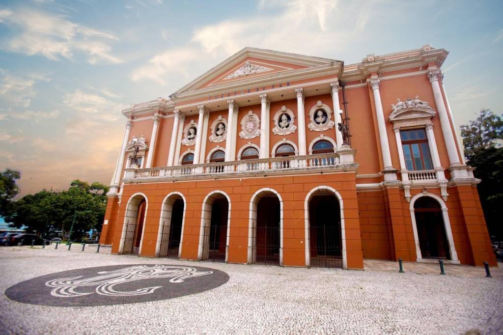 Fachada do Teatro da Paz em Belém, Pará, com paredes cor de salmão e detalhes brancos. O edifício tem um frontão triangular, colunas coríntias, bustos em nichos e arcos no térreo. O piso em frente é de pedras claras com um mosaico escuro no centro, sob um céu azul com nuvens esparsas