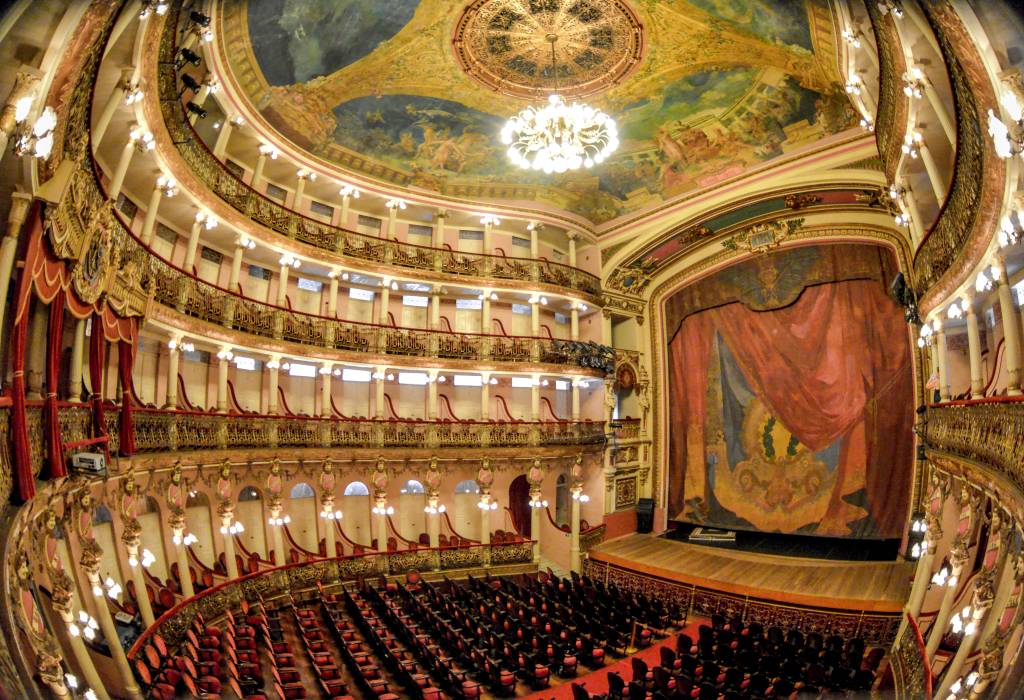Interior de um teatro opulento com poltronas vermelhas e cinco andares de camarotes dourados e rosados. Um lustre central ilumina o teto pintado com cenas mitológicas. O palco tem uma cortina vermelha e azul com detalhes dourados. A imagem é capturada com uma lente olho de peixe, distorcendo as laterais.