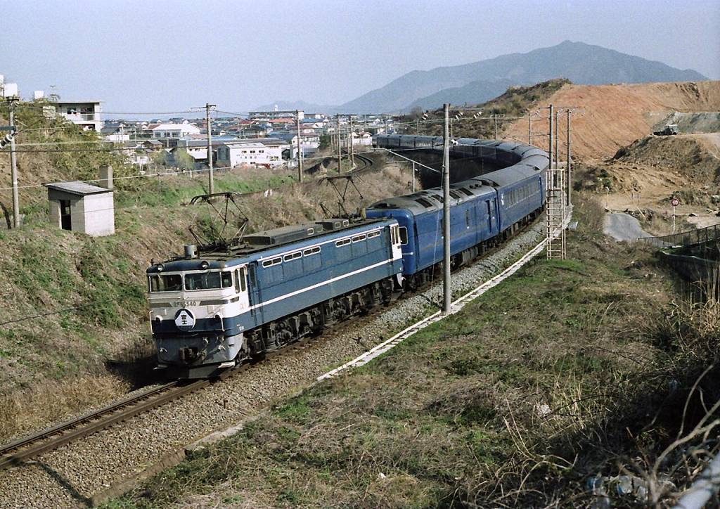Trem azul e branco em curva acentuada, com vagões azuis, em trilhos de cascalho. Ao fundo, casas e montanhas, sob céu claro.