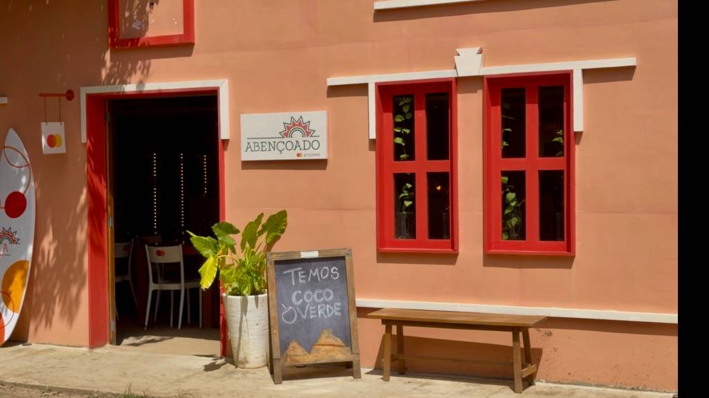 Fachada de um estabelecimento com paredes cor de pêssego e detalhes em vermelho vibrante. À esquerda, uma prancha de surfe branca com detalhes coloridos. A porta de entrada vermelha revela um interior escuro com cadeiras brancas. Acima da porta, uma placa branca com o nome ABENÇOADO. Duas janelas vermelhas com plantas verdes dentro. Em frente, um vaso branco com planta e um cavalete de lousa preta com a frase TEMOS COCO VERDE. À direita, um banco de madeira. A luz do sol cria sombras na parede