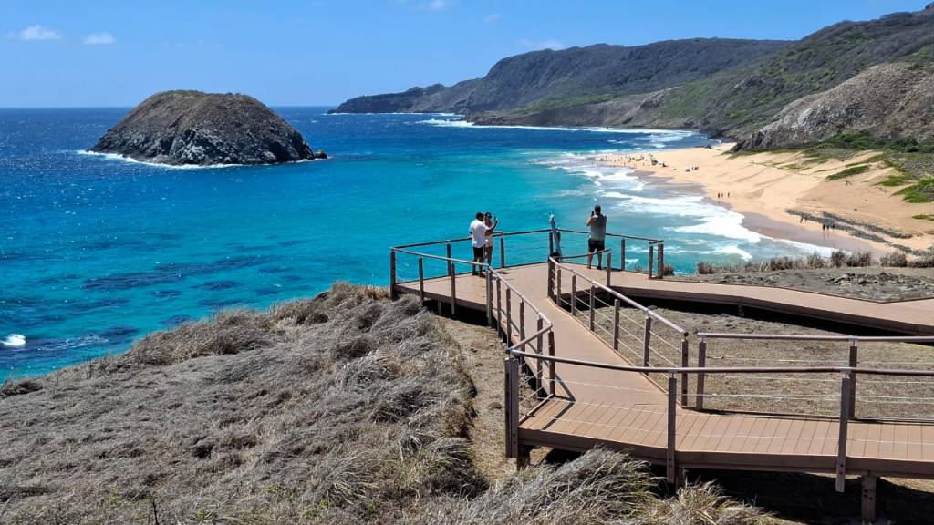 Vista aérea de uma praia paradisíaca com mar azul-turquesa e areia dourada. No primeiro plano, uma passarela de madeira com corrimão metálico serpenteia por uma encosta coberta de vegetação seca. Três pessoas observam a paisagem do mirante, uma delas tirando foto. Ao fundo, uma ilha rochosa emerge do oceano e montanhas verdes emolduram a costa. O céu está claro e azul