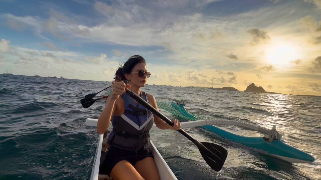 Mulher de óculos escuros e colete salva-vidas cinza e azul rema em canoa no mar. O sol se põe no horizonte, iluminando o céu com tons alaranjados e nuvens esparsas. Ao fundo, ilhas e barcos pontilham a paisagem marítima