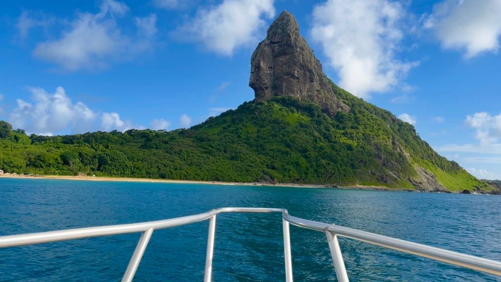 Vista da proa de um barco, com o Morro do Pico em Fernando de Noronha ao fundo. O morro é uma formação rochosa escura e pontiaguda, coberta por vegetação verde exuberante. O mar azul-turquesa ocupa o primeiro plano, com a costa arenosa e mais vegetação à esquerda. O céu azul claro tem nuvens brancas esparsas