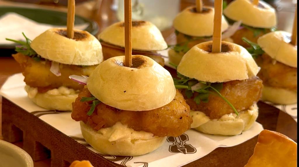 Mini sanduíches com pão redondo, recheio de peixe empanado, molho branco, cebola roxa e folhas verdes, espetados com palitos de madeira, servidos em uma bandeja de madeira escura com guardanapos brancos