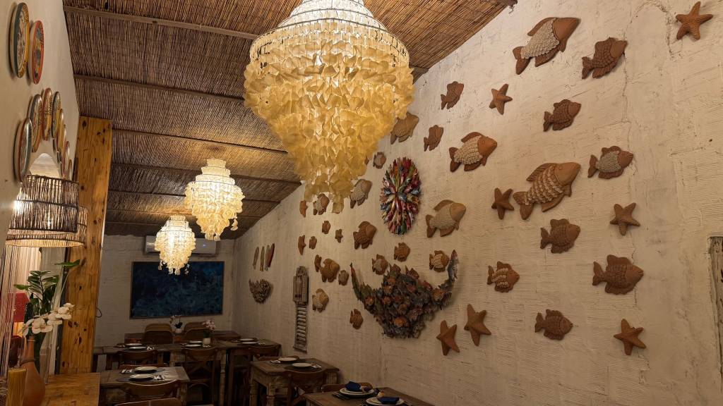 Ambiente de restaurante com teto de palha, lustres de conchas e paredes brancas decoradas com peixes e estrelas do mar de cerâmica, mesas de madeira com pratos e talheres