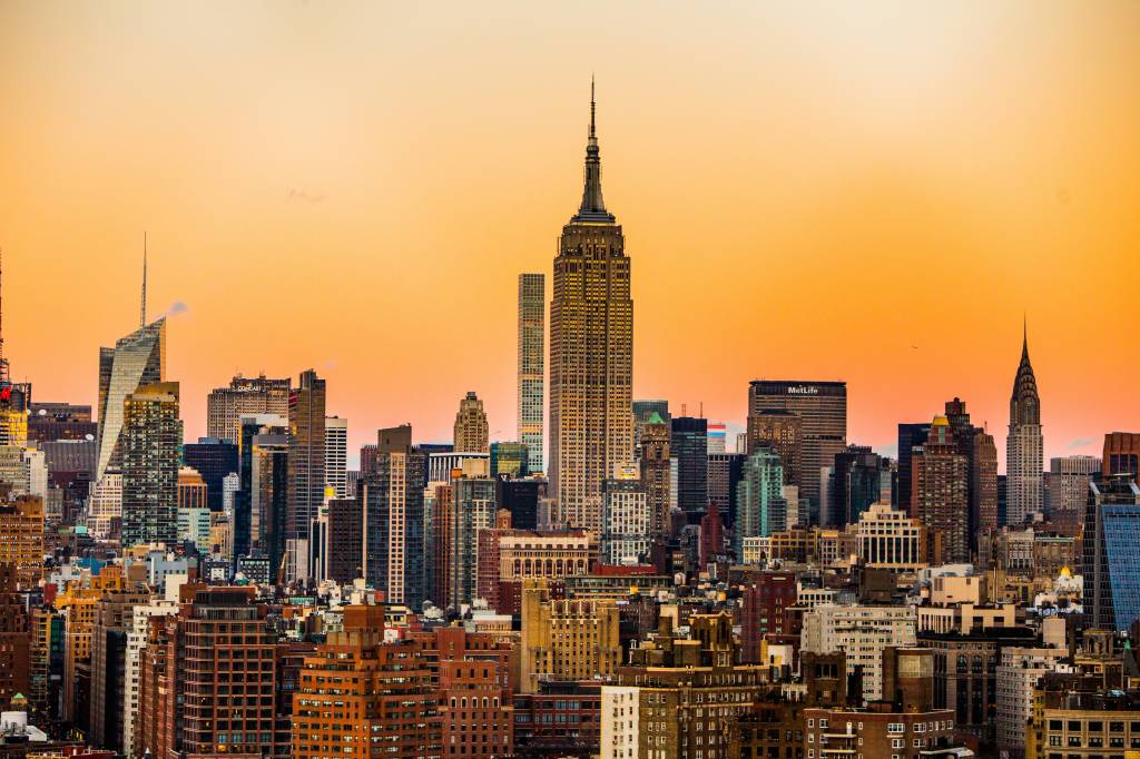 Vista panorâmica da cidade de Nova York ao pôr do sol, com o Empire State Building e o Chrysler Building se destacando entre os arranha-céus, sob um céu laranja vibrante