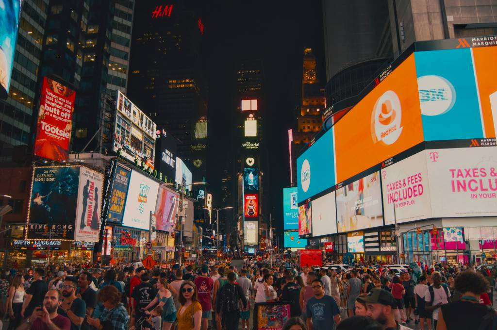 Multidão de pessoas em Times Square, Nova York, à noite, com edifícios altos e inúmeros painéis digitais coloridos exibindo anúncios luminosos de marcas e espetáculos, criando uma atmosfera vibrante e movimentada