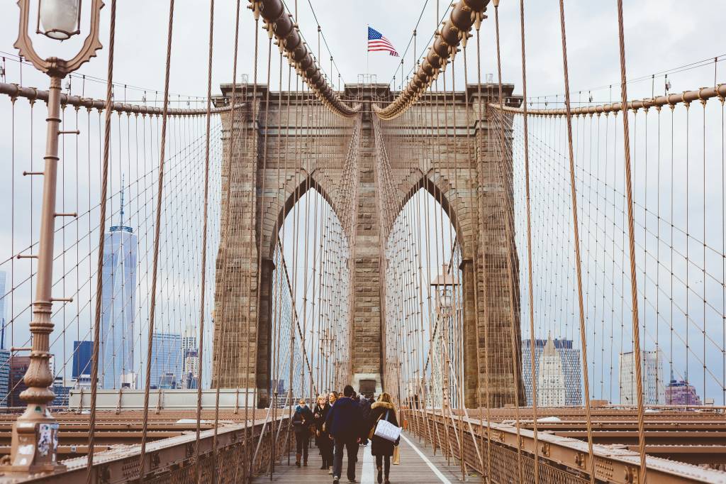 Pessoas caminhando sobre a Ponte do Brooklyn, com suas torres de pedra e cabos de aço. Uma bandeira dos EUA tremula no topo da torre central. Ao fundo, arranha-céus de Nova York sob um céu nublado