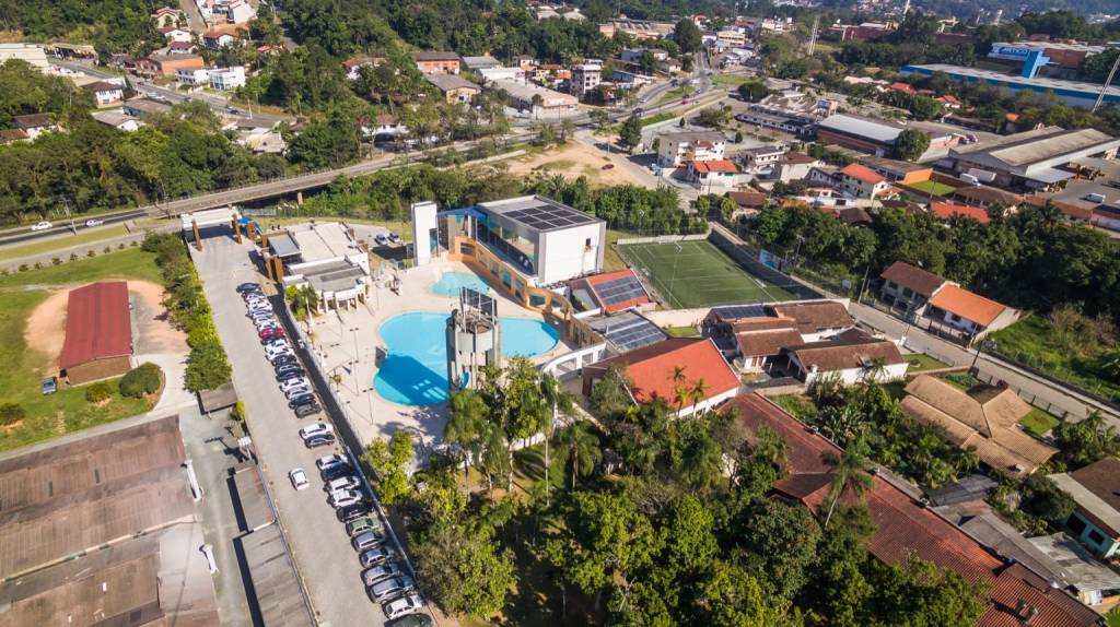 Vista aérea de um complexo de lazer com piscinas azuis, um prédio moderno com painéis solares no telhado e um campo de futebol gramado. Ao redor, há um estacionamento com carros, casas com telhados vermelhos e muitas árvores verdes, com uma rodovia e mais construções ao fundo